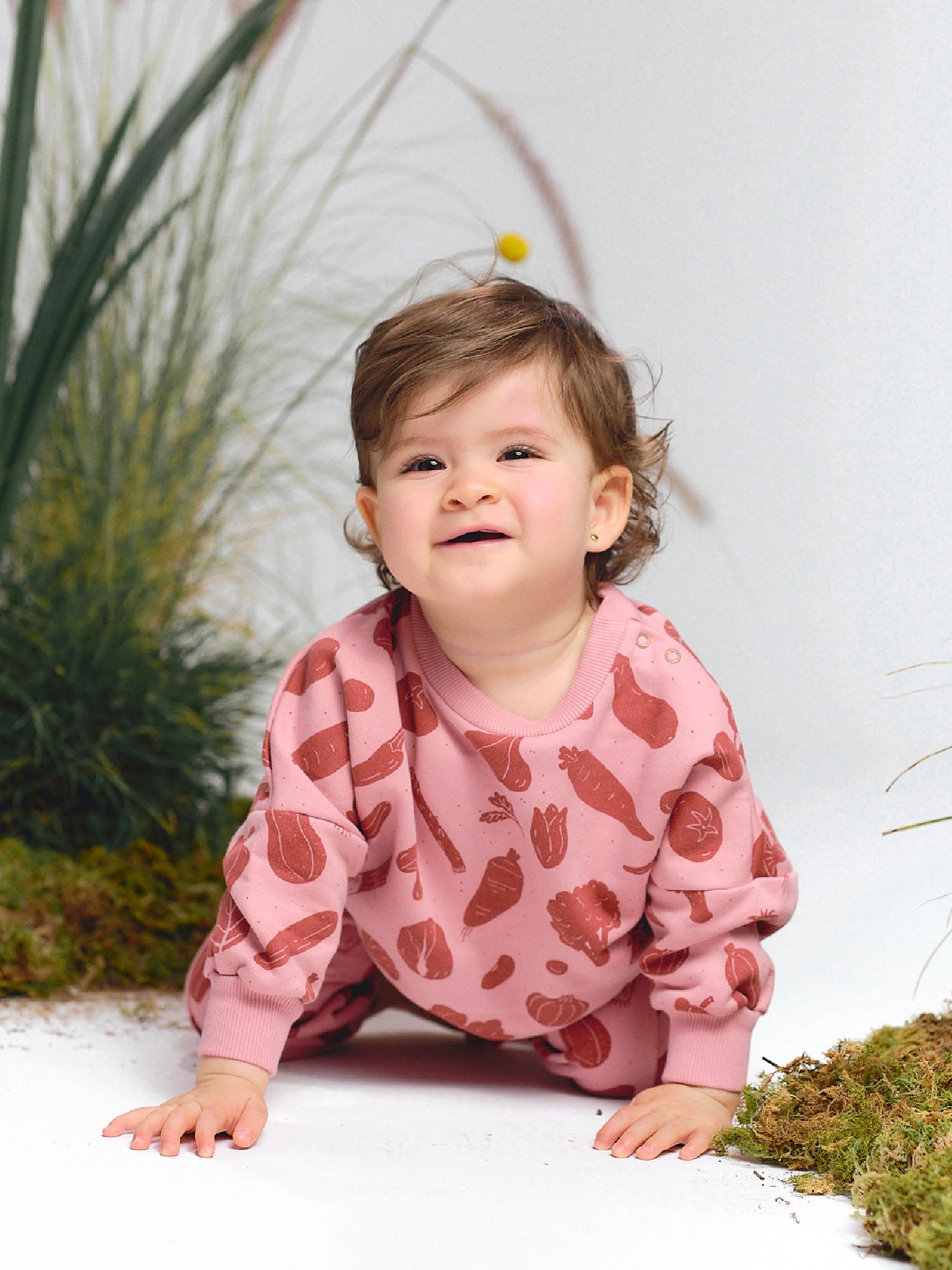 Baby crawling on the floor in a pink sweatshirt with vegetable prints in red tones, smiling.