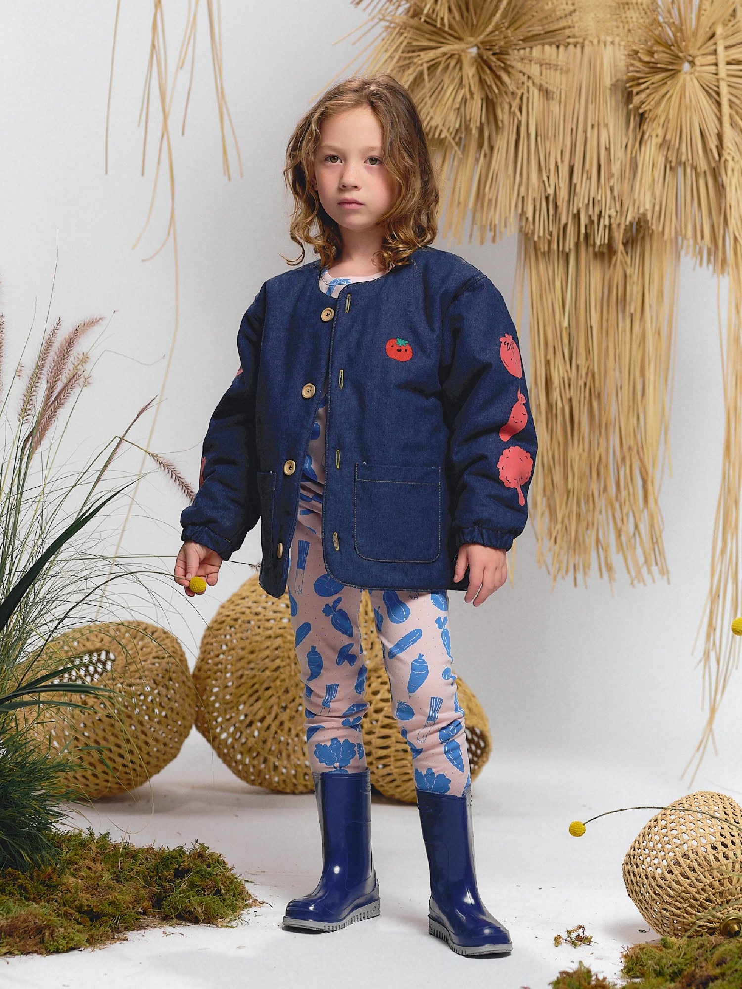 Girl wearing pink leggings with Veggie Fiesta vegetable print and light blue T-shirt underneath a blue denim jacket embroidered with red tomatoes. Styled with navy rain boots.