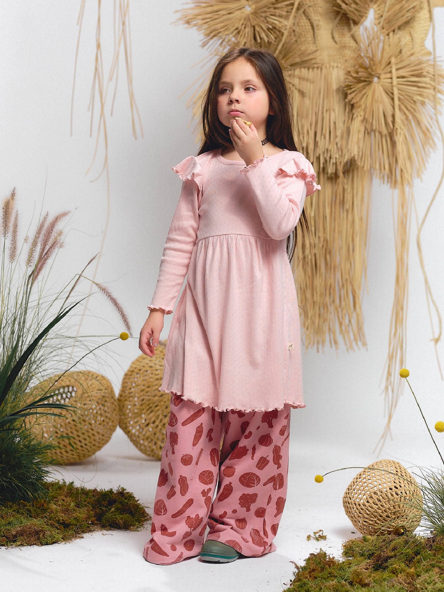 Girl wearing pale pink long-sleeve dress with ruffle shoulders, paired with wide-leg pants featuring Veggie Fiesta vegetable print. Styled with green shoes.