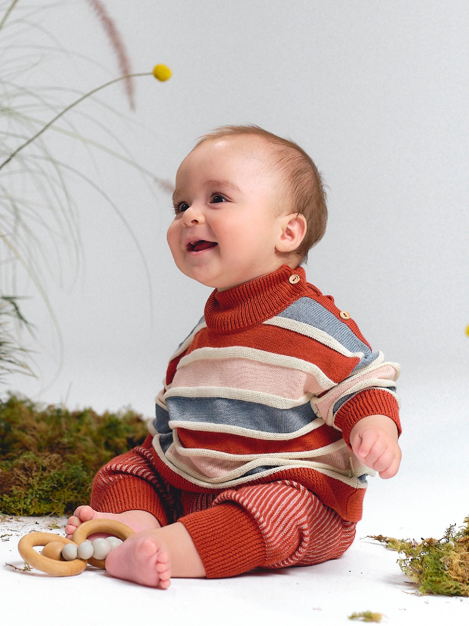 Baby sitting and smiling, dressed in a striped knit sweater in brick red, pink and blue with matching knit pants.
