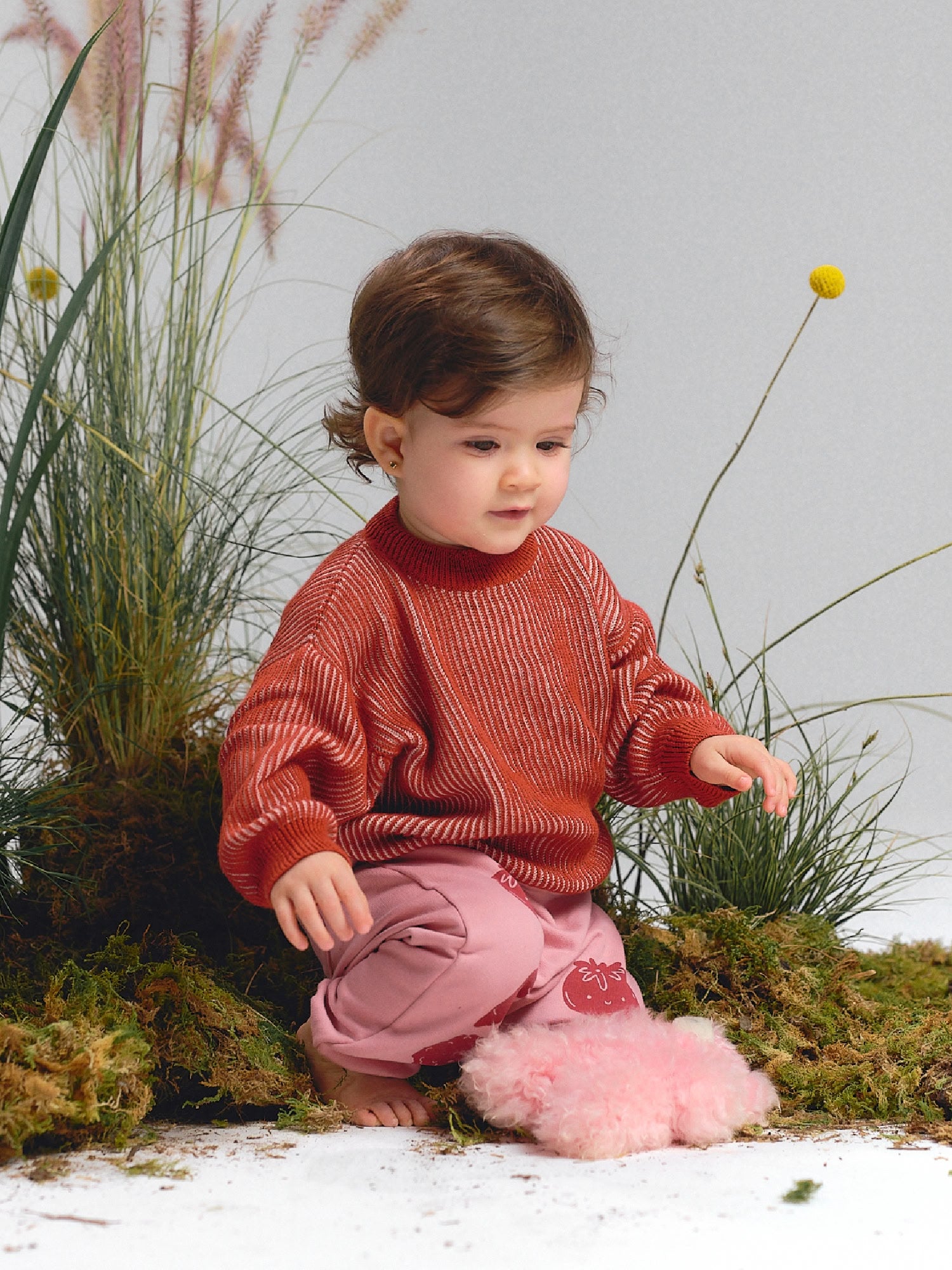Baby girl wearing a red knit sweater and pink pants with tomato prints, playing with a pink plush toy.