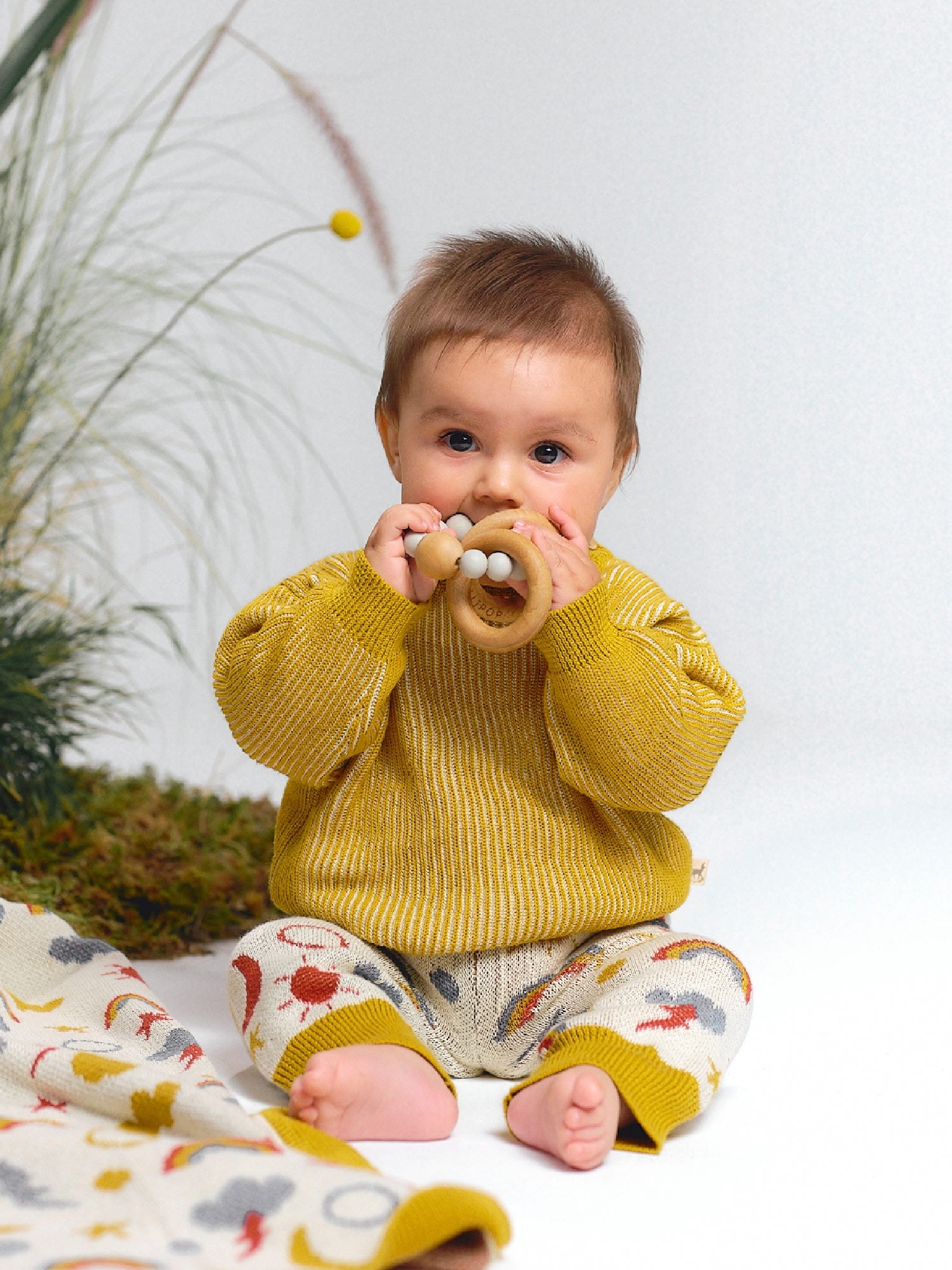 Baby in a yellow knit sweater and jacquard knit pants with colorful sky motifs, holding a wooden rattle.