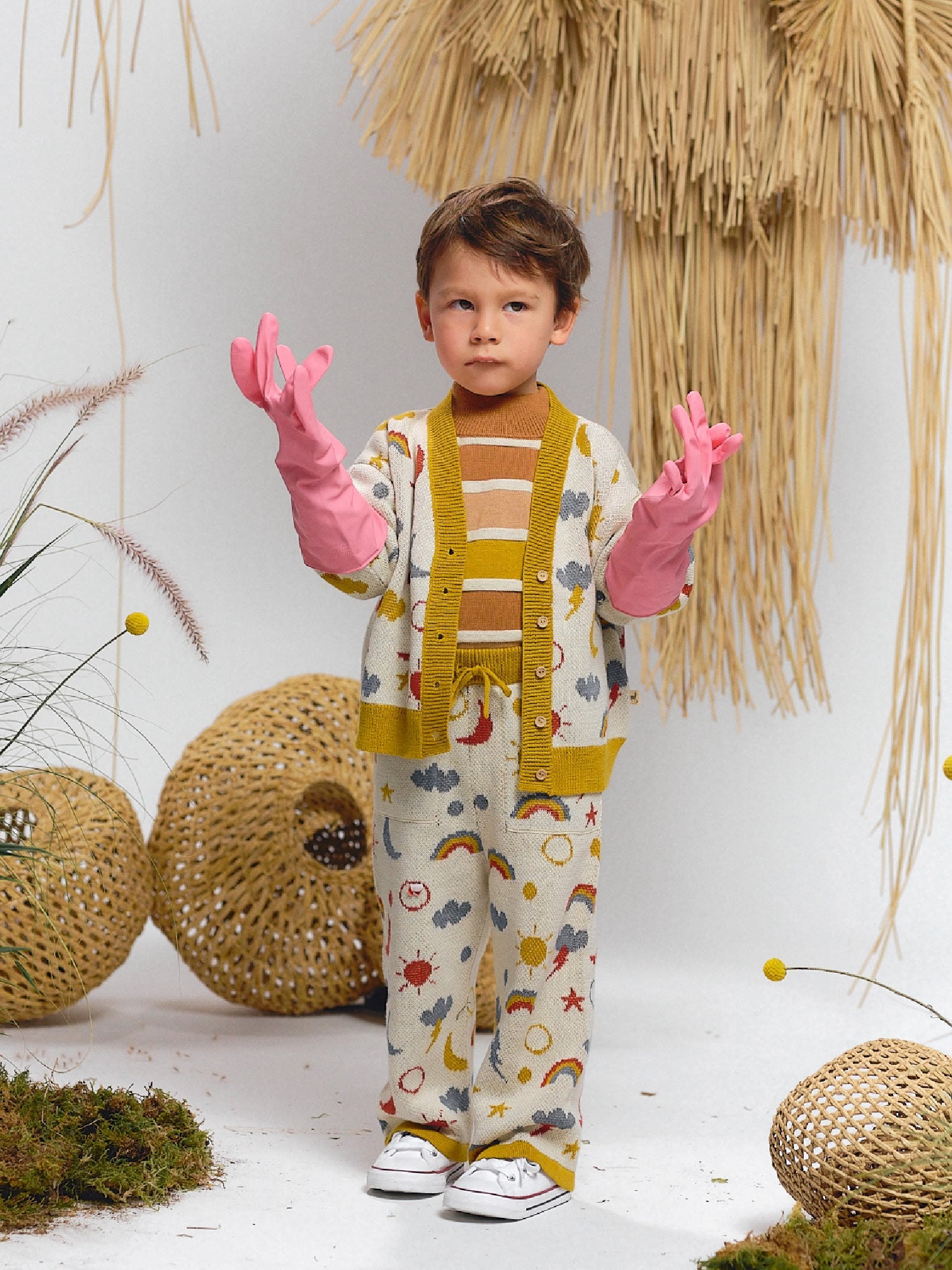 Boy wearing a rainbow-themed knit cardigan and pants set with sun, moon, and stars motifs, styled with a striped sweater underneath, pink gloves, and white sneakers.