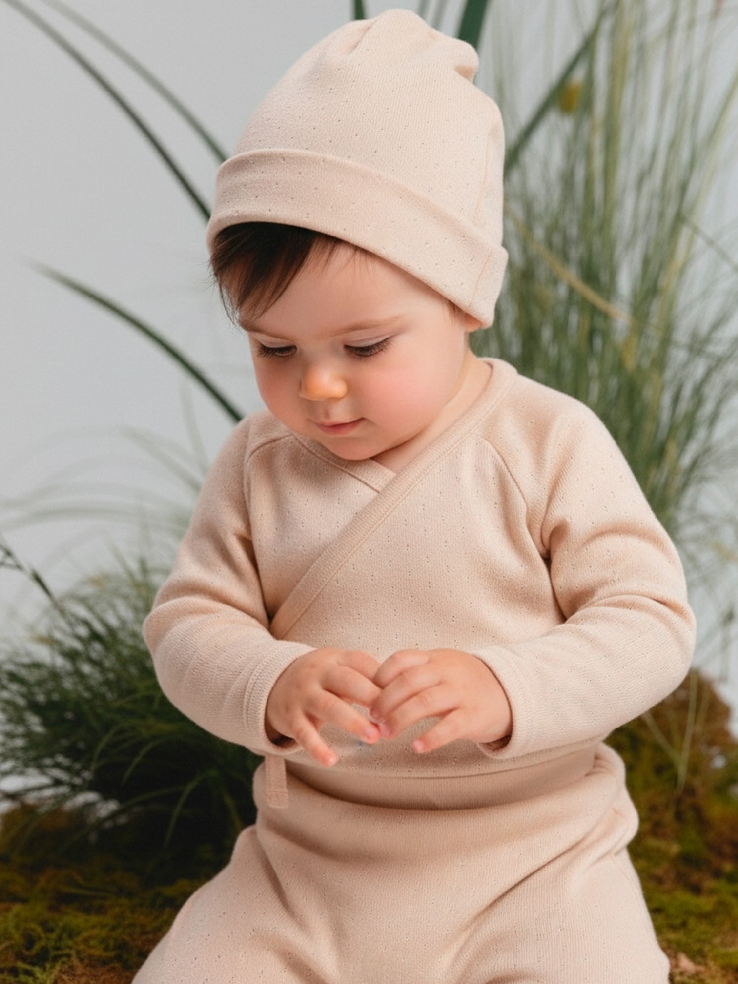 Baby wearing a soft pink kimono set with wrap-style top and matching pants, styled with a printed hat, sitting on a natural moss background.