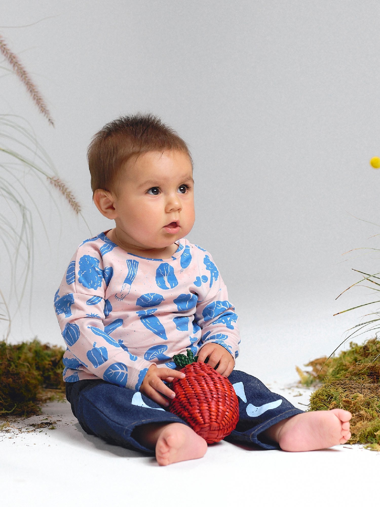 Baby sitting on the floor wearing a pink t-shirt with blue vegetable prints and denim pants with blue sky motifs, holding a woven red strawberry toy.
