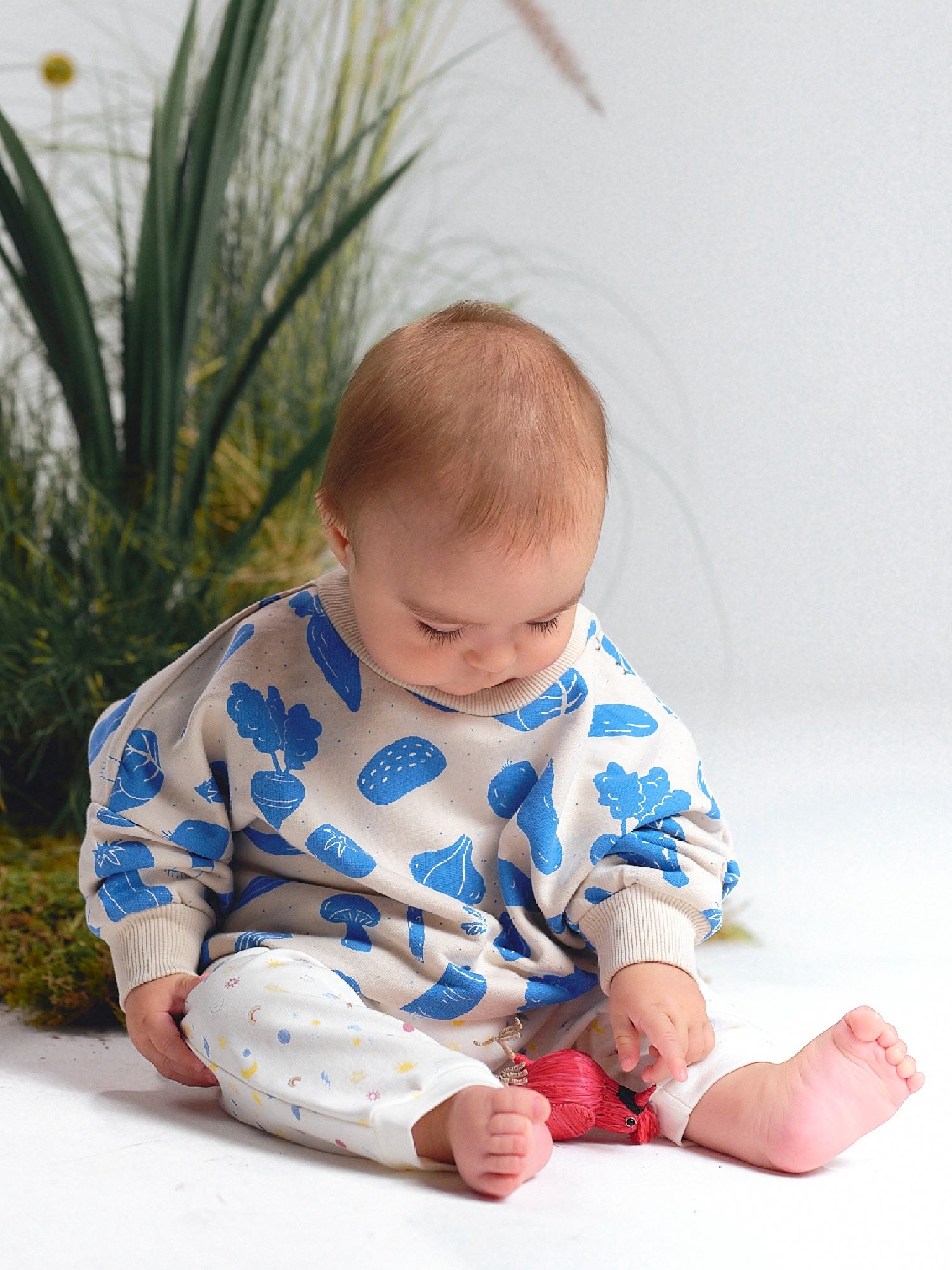 Baby looking down while wearing a cream sweatshirt with blue vegetable prints and white pants with small sky motifs.