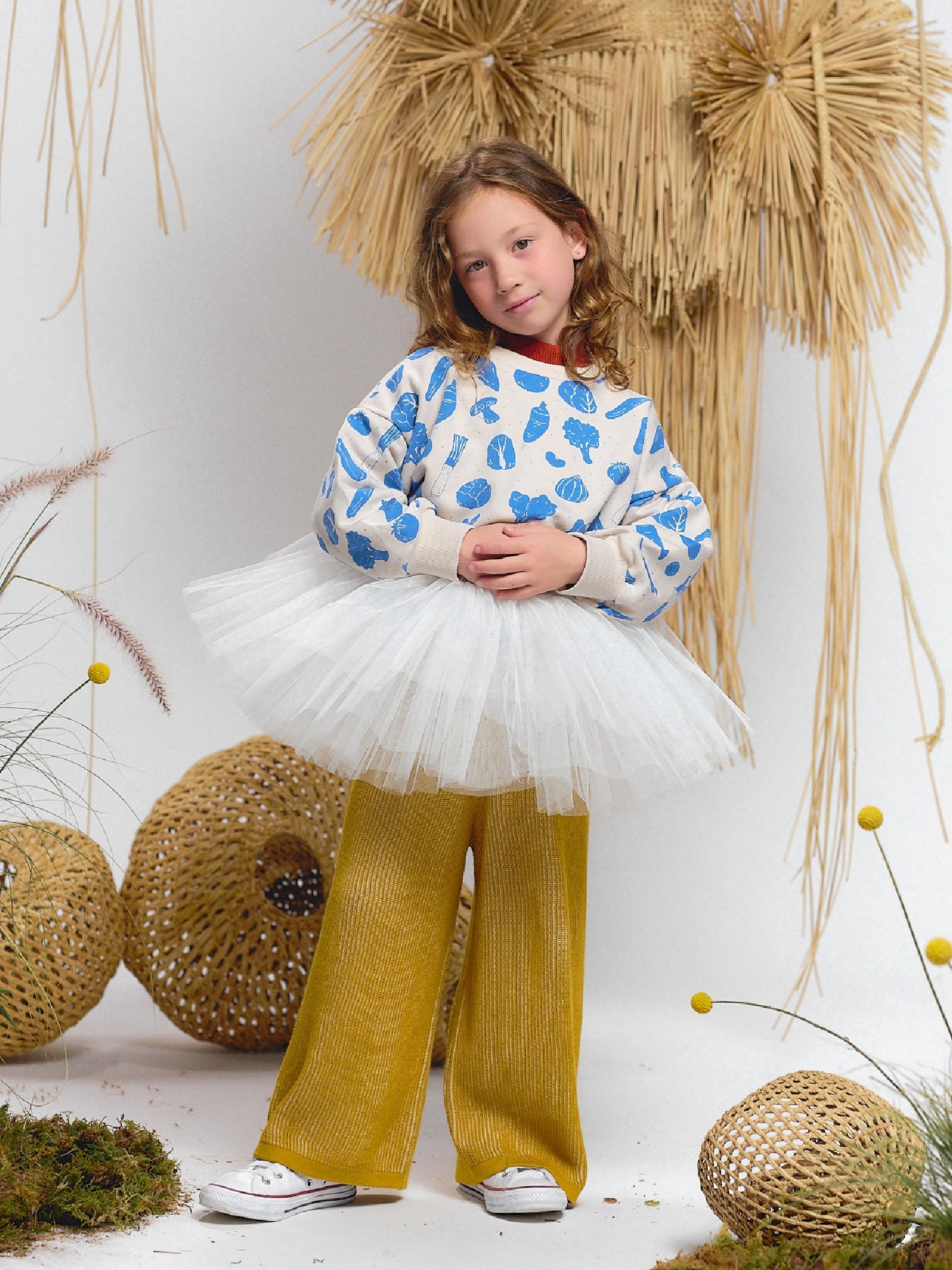 Girl in a white sweatshirt with blue veggie fiesta print, paired with wide mustard knit pants, white tutu skirt overlay, and white sneakers.