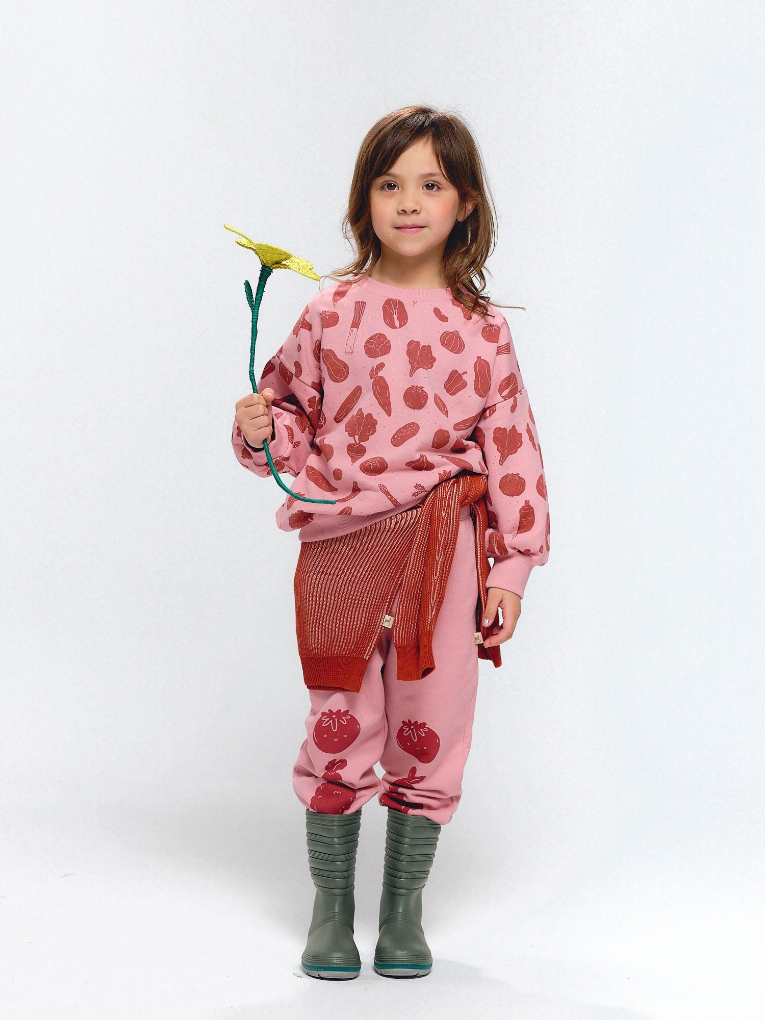 Girl wearing oversized pink sweatshirt and joggers with Veggie Fiesta vegetable print. Outfit styled with a red knit sweater tied at the waist and green boots. Holding a yellow flower.
