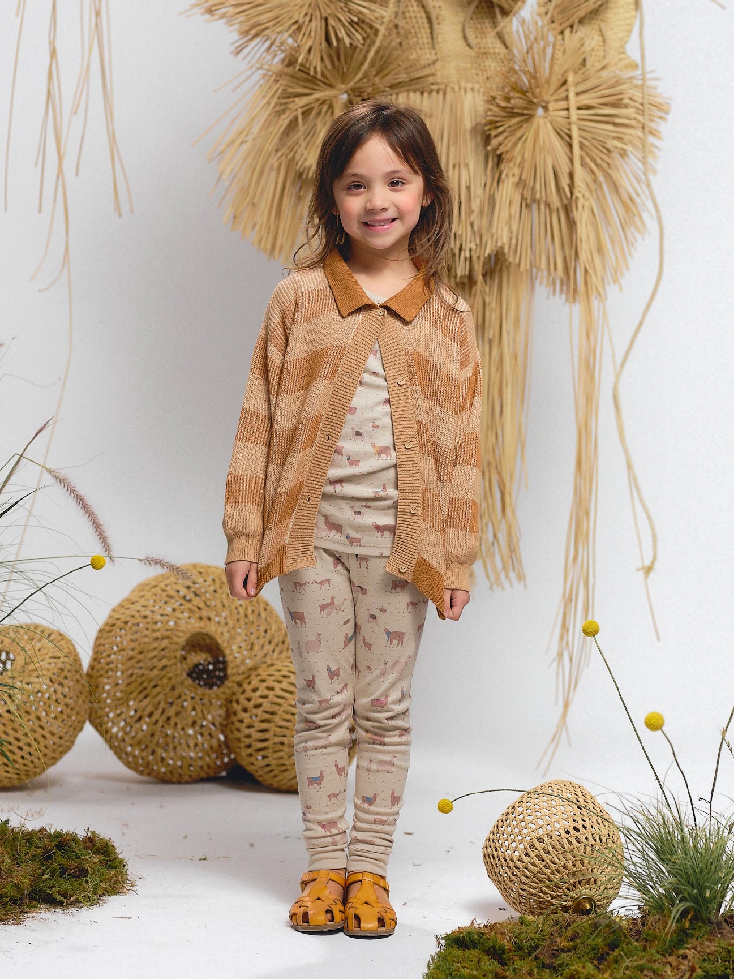 Alt: Smiling girl wearing a beige alpaca print long-sleeve top and matching leggings, styled with a striped knit cardigan and mustard sandals.