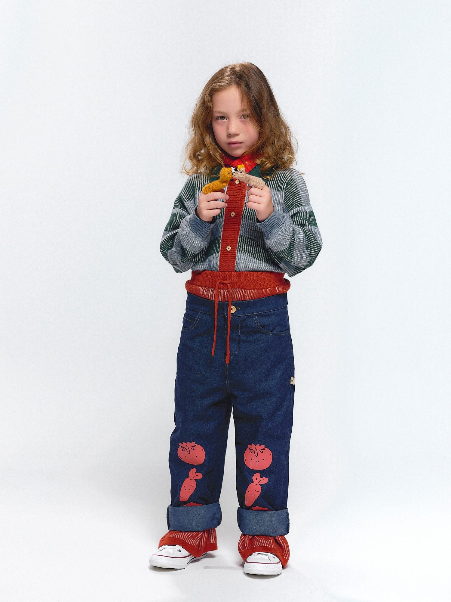 Girl in denim carrot, tomato-print pants with rolled cuffs, green striped shirt, red knit scarf, and red leg warmers, holding a toy animal.