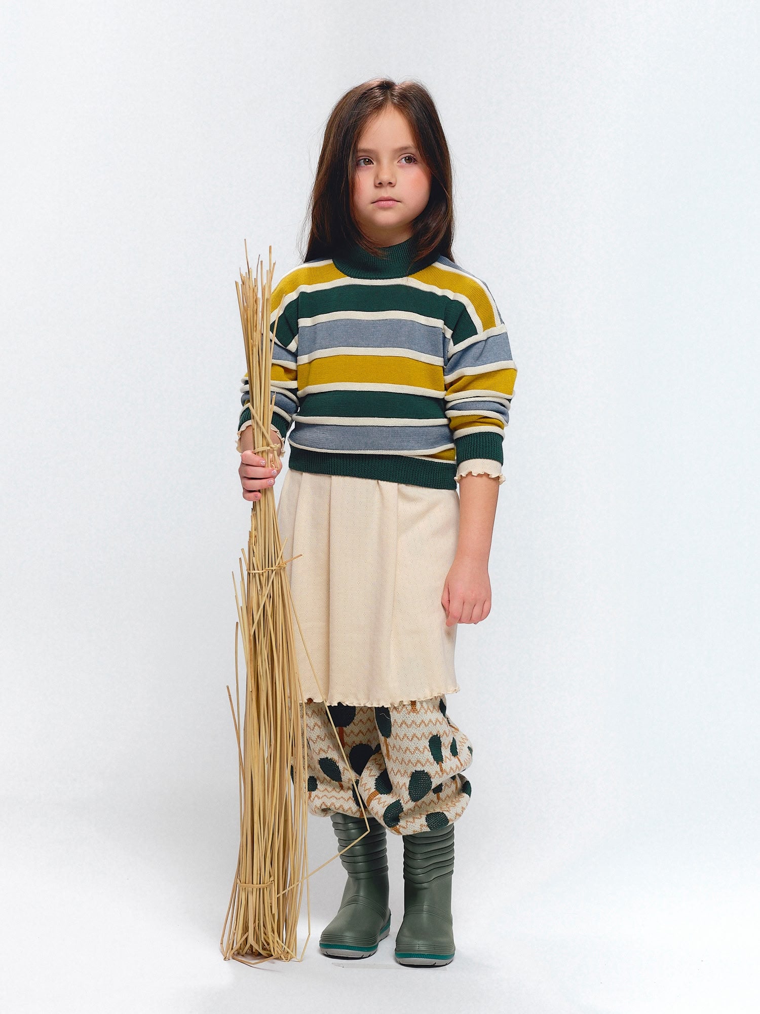 Girl wearing striped knit sweater in green, blue, and yellow, layered over a cream skirt with forest jacquard knit pants and green boots, holding straw sticks.