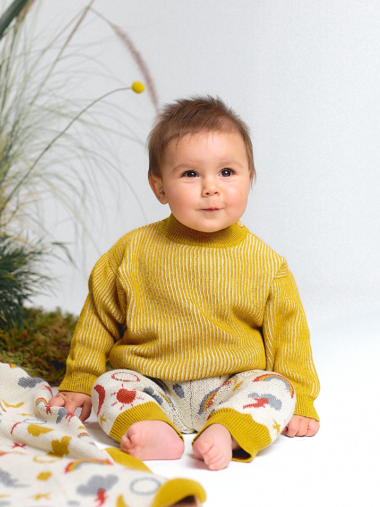 Baby in a yellow knit sweater and sky motif jacquard knit pants, sitting on the floor with a matching blanket.