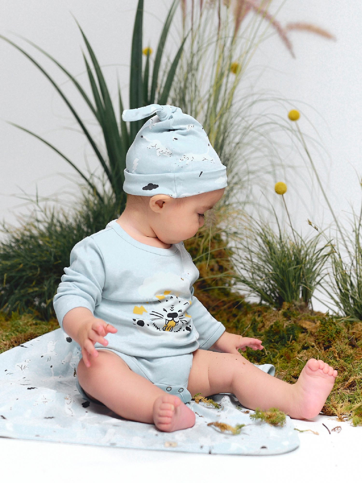 Baby sitting on a blanket wearing a light blue onesie and hat with thunder puma print, looking down.
