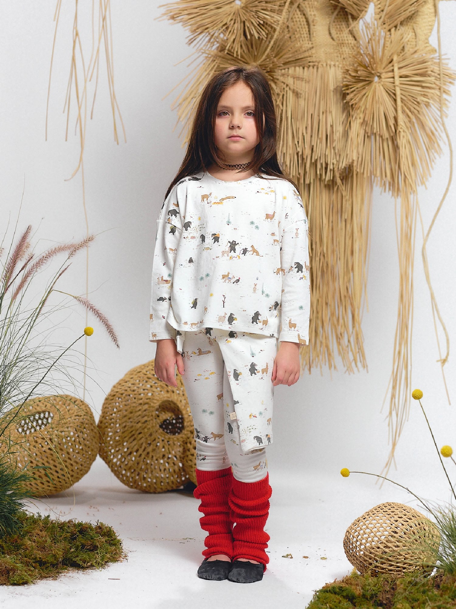 Girl wearing an ivory long-sleeve top and leggings featuring Andean animals print — spectacled bear, alpaca, puma, and native fauna — styled with red leg warmers and black flats.