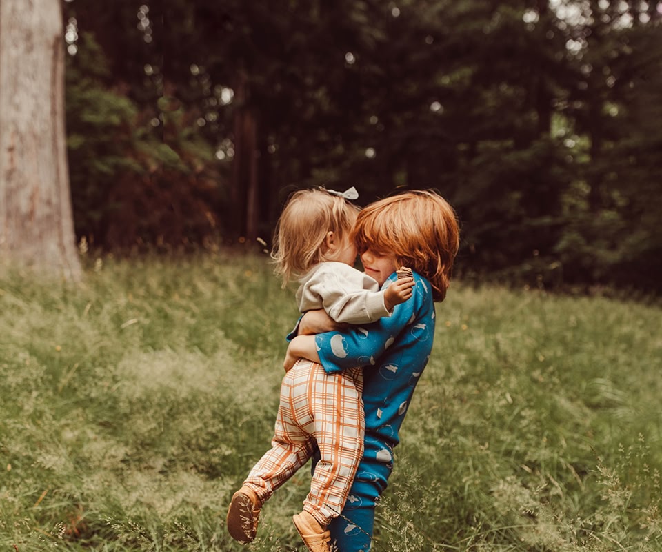 Two kids playing outdoors wearing colorful garments from past Red Caribou collections. Banner for the Sale section with discounts on organic clothing for babies and kids.